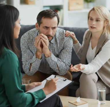 Couple in therapy session, man appears concerned while woman offers support; therapist taking notes on clipboard